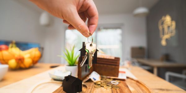 Hand holding house keys above wooden table with coins and house model.