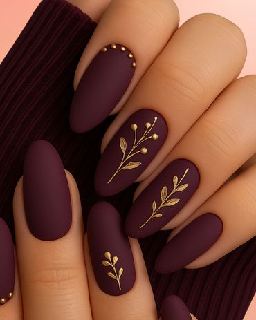 Matte burgundy nails with gold floral designs and studs.