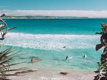 Surfers waiting on bright turquoise waves near a rocky beach under a clear sky.