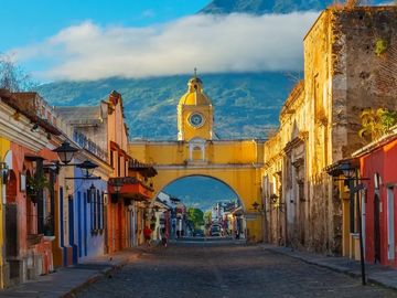 Historic yellow clock tower arch over a cobblestone street with mountains in the background.