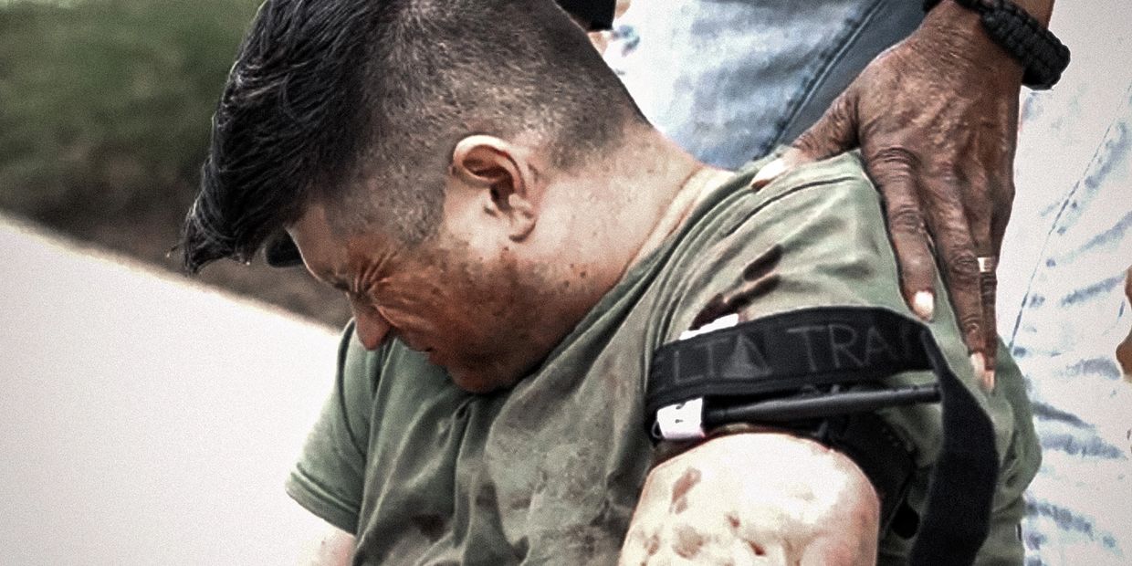 A man in pain with a tourniquet on his arm, being comforted by another person.