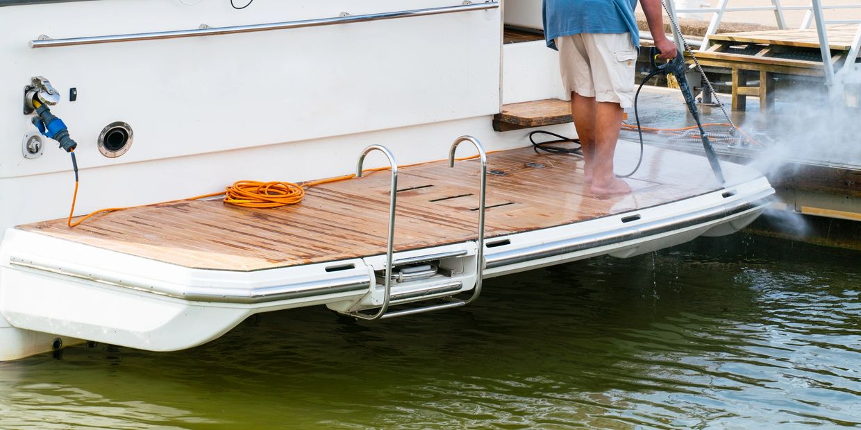 Person pressure washing a boat's wooden deck near the water.