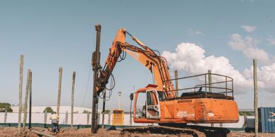 Orange excavator at a construction site with wooden poles and a worker.
