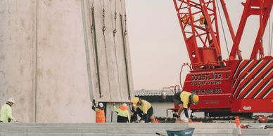 Construction workers guide a large concrete panel lifted by a red crane at a building site.