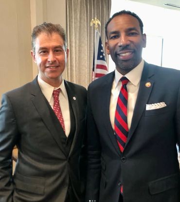 Two men in suits smiling, standing indoors with an American flag in the background.