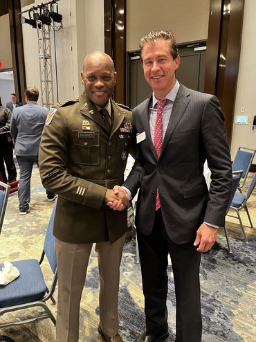 Two men shaking hands, one in military uniform, the other in a suit, smiling at an indoor event.