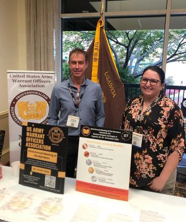 Two representatives at a US Army Warrant Officers Association booth with informational displays.