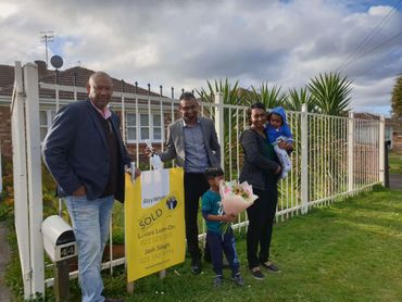 Family celebrates buying a new home with a sold sign and flowers.