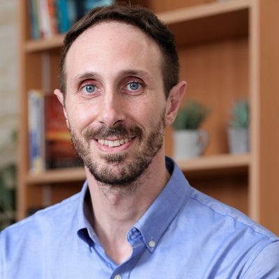 Smiling man in blue shirt with bookshelf background.