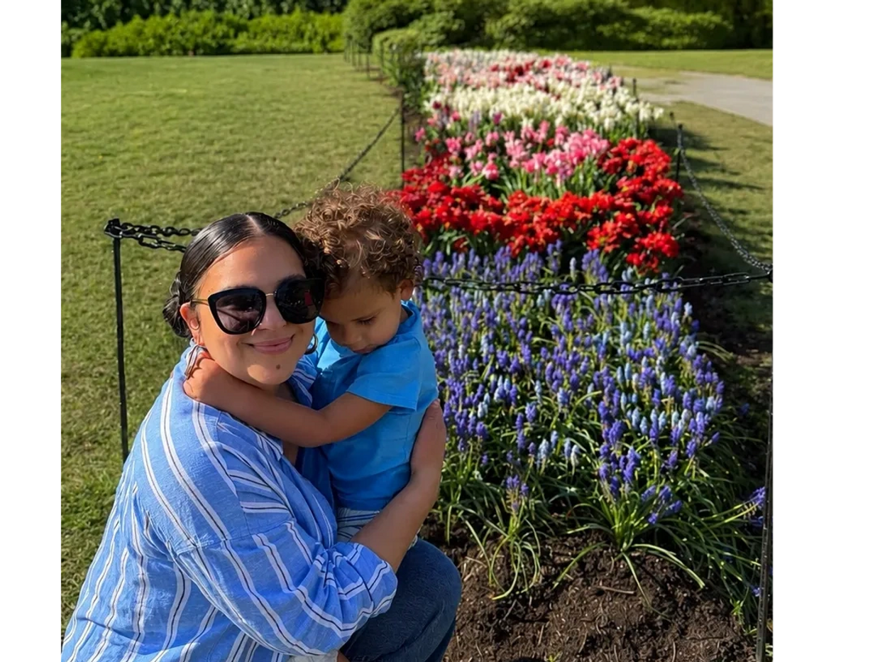 A woman in sunglasses hugs a child near a colorful flower bed in a park.