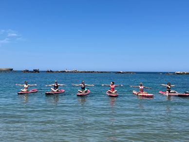 Six people practicing paddleboarding yoga on calm ocean waters under a clear blue sky.