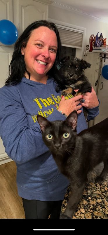 Smiling woman holding a small dog with a black cat in the foreground.
