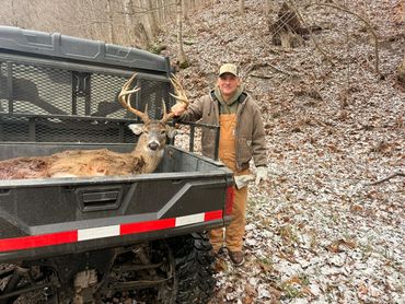 Hunter stands beside a large deer in the back of an ATV in a snowy forest.