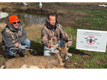 Two men pose with a large deer and a drone by a pond, next to a business sign.