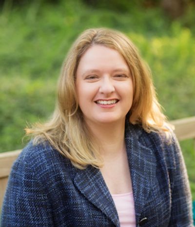 Michelle E. Watson on a park bench in a blue suit with pink top. Blonde with with a broad smile.