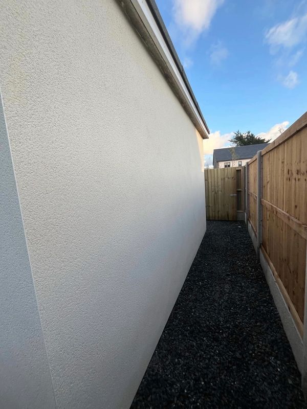 Narrow outdoor pathway between a white wall and wooden fence under a blue sky.