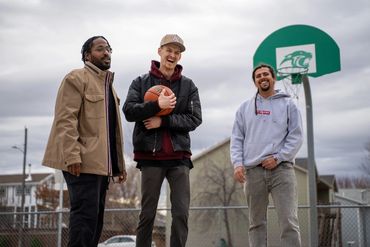 Three friends smiling on an outdoor basketball court with a basketball hoop in the background.