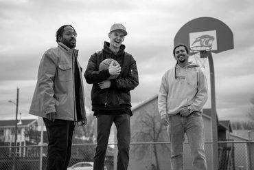 Three friends smiling on an outdoor basketball court with a basketball hoop in the background.