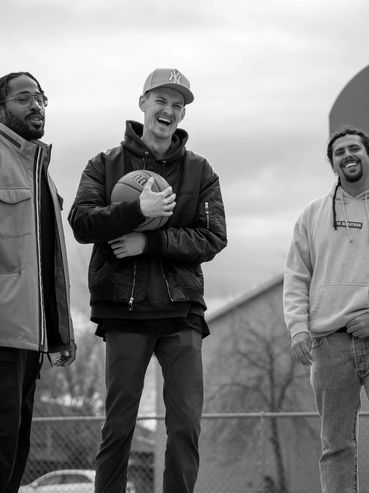 Three friends smiling on an outdoor basketball court with a basketball hoop in the background.