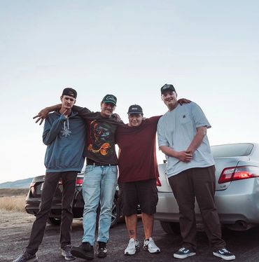 Four friends posing together outdoors beside two cars during sunset.