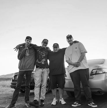 Four friends posing together outdoors beside two cars during sunset.