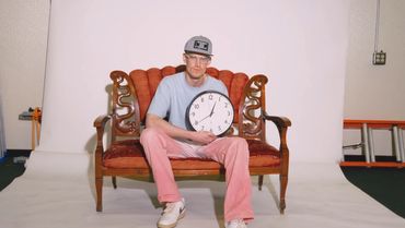 Man sitting on vintage couch holding a clock showing 12:06.