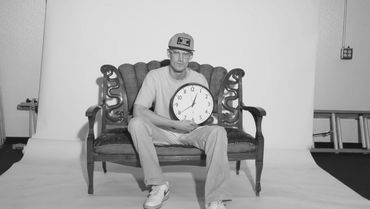 Man sitting on vintage couch holding a clock showing 12:06.