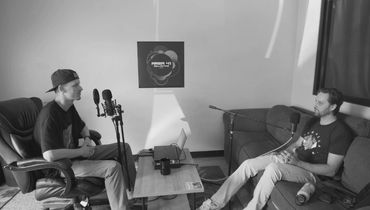 Two men recording a podcast in a casual room setup with microphones and a laptop.