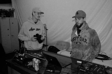 Two men standing by a DJ setup with laptops and sound equipment.