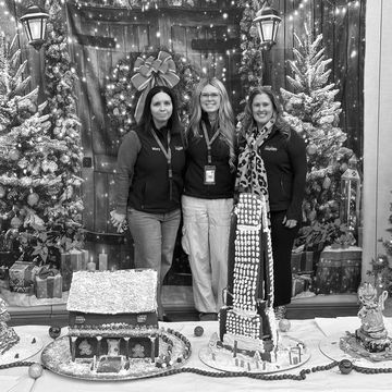 Three women posing with gingerbread houses in festive winter backdrop.