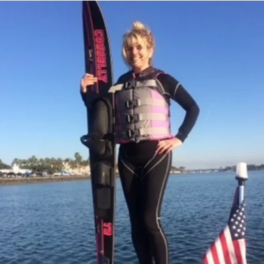 Woman in wetsuit holding a water ski near a body of water with an American flag.