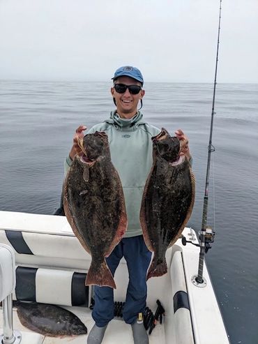 Man proudly holds two large flatfish on a boat in calm sea.