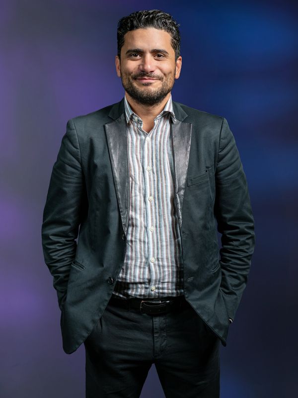 Man with dark hair and beard in a black blazer and striped shirt smiling confidently.
