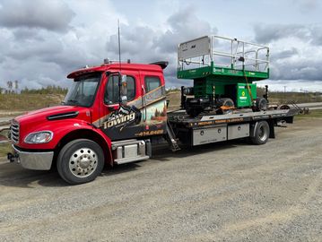 Red towing truck carrying green lift equipment on flatbed under cloudy sky.