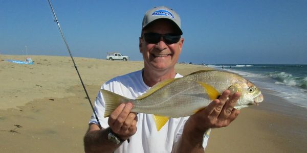 Man smiling and holding a fish on a sandy beach with ocean waves.