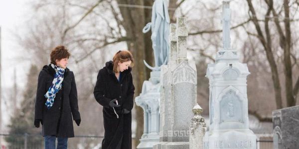 Two women in black coats visiting a snowy cemetery.
