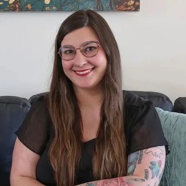 Smiling woman with glasses and a colorful floral tattoo on her arm sitting on a couch.