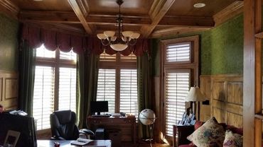 Cozy home office with wooden paneling, green curtains, and a globe near large shuttered windows.