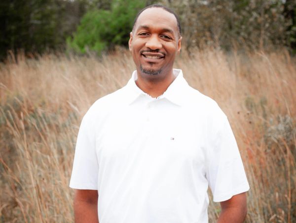 Smiling man in a white polo shirt standing in a field of tall grass.