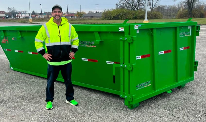 Man in neon yellow hoodie standing by a large green dumpster.