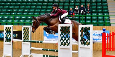 Equestrian rider jumping a horse over an obstacle in an indoor arena.