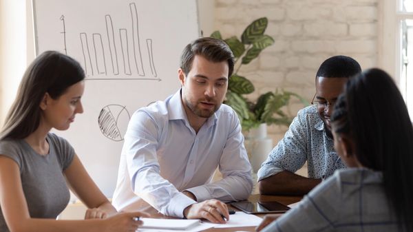 A group of professionals engaged in a meeting around a table, with a whiteboard displaying graphs