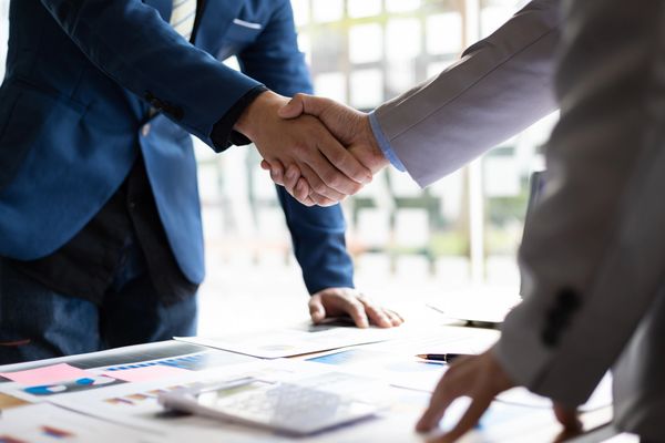 Two business professionals in suits shake hands over a table covered with documents and charts