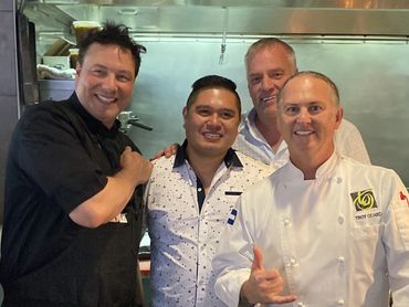 Four men posing and smiling in a kitchen, one wearing a chef's coat.