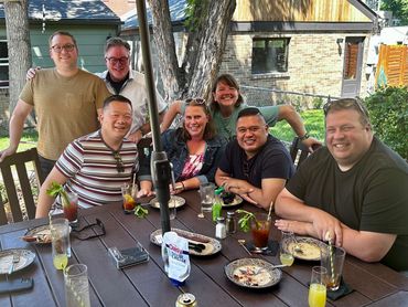 Group of friends smiling outdoors at a table with drinks and food.