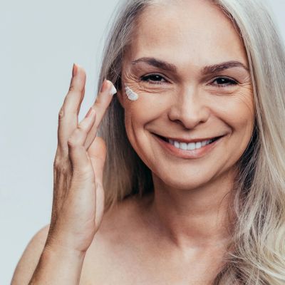 an image of an older woman smiling, applying cream on her face