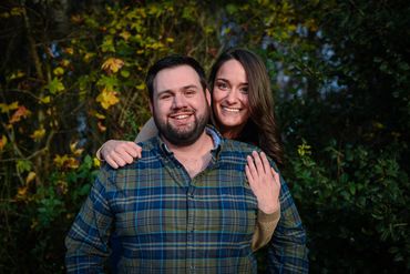 Smiling couple embracing outdoors with autumn foliage behind.