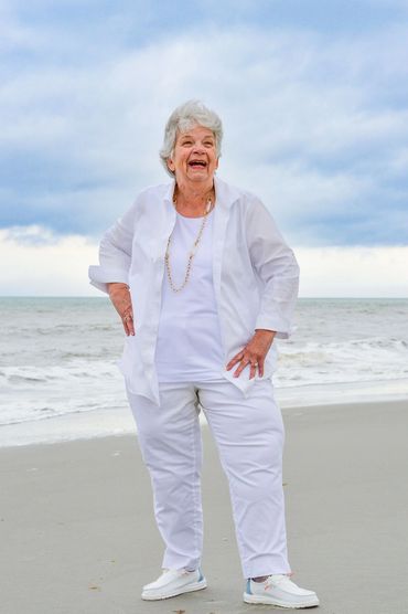 Elderly woman joyfully posing on the beach in an all-white outfit.