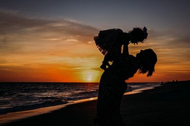 A mother lifts her child at sunset on the beach, silhouetted against the colorful sky.