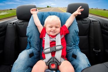 Happy toddler with arms raised in a car seat inside a convertible on a sunny day.